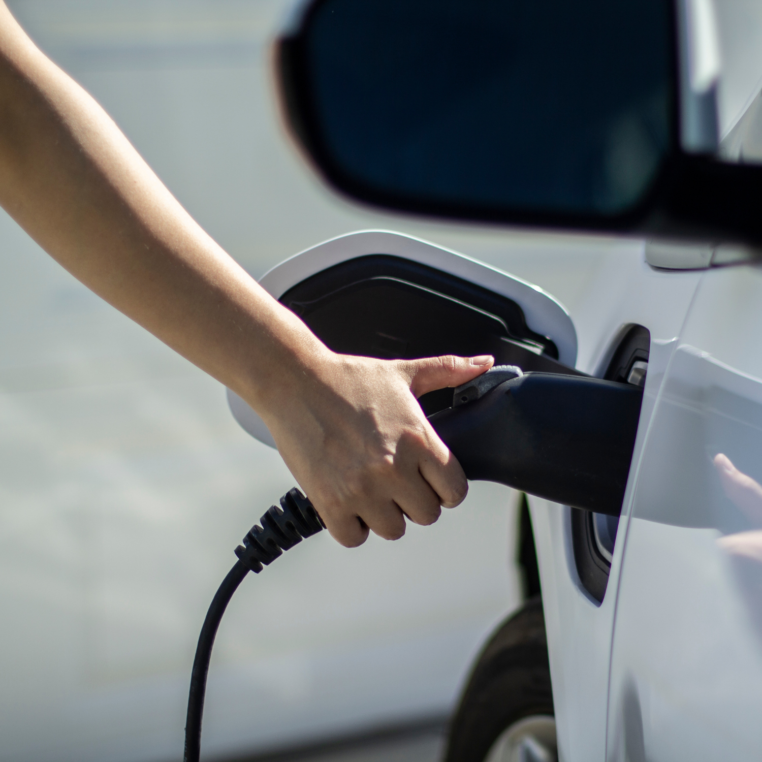 Mano enchufando cargador a un vehículo eléctrico blanco, en un entorno urbano, representando uso cotidiano de movilidad eléctrica.