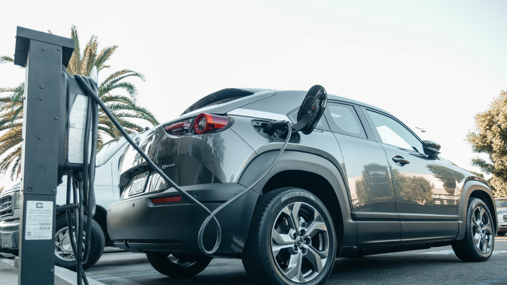 Vehículo SUV eléctrico de color gris conectado a estación de carga pública en entorno urbano, con árboles y palmeras de fondo, reflejando movilidad sostenible y tecnología limpia.