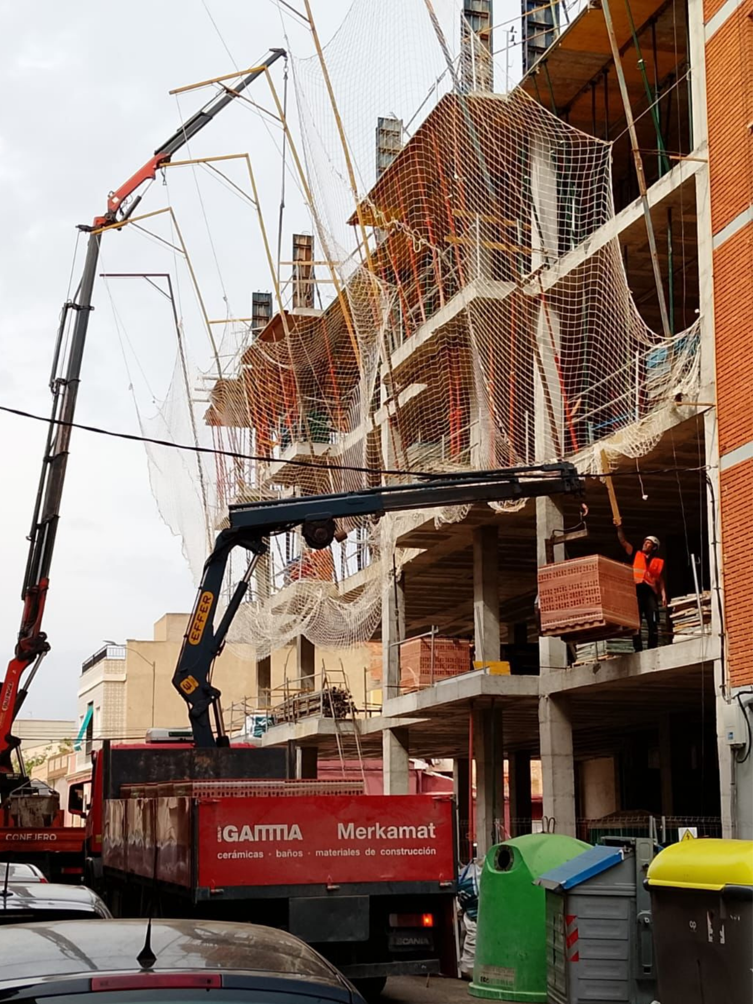 Grúa hidráulica elevando palets de ladrillos en una construcción de edificio con redes de seguridad instaladas y operarios en obra, frente a camión de suministro de materiales.