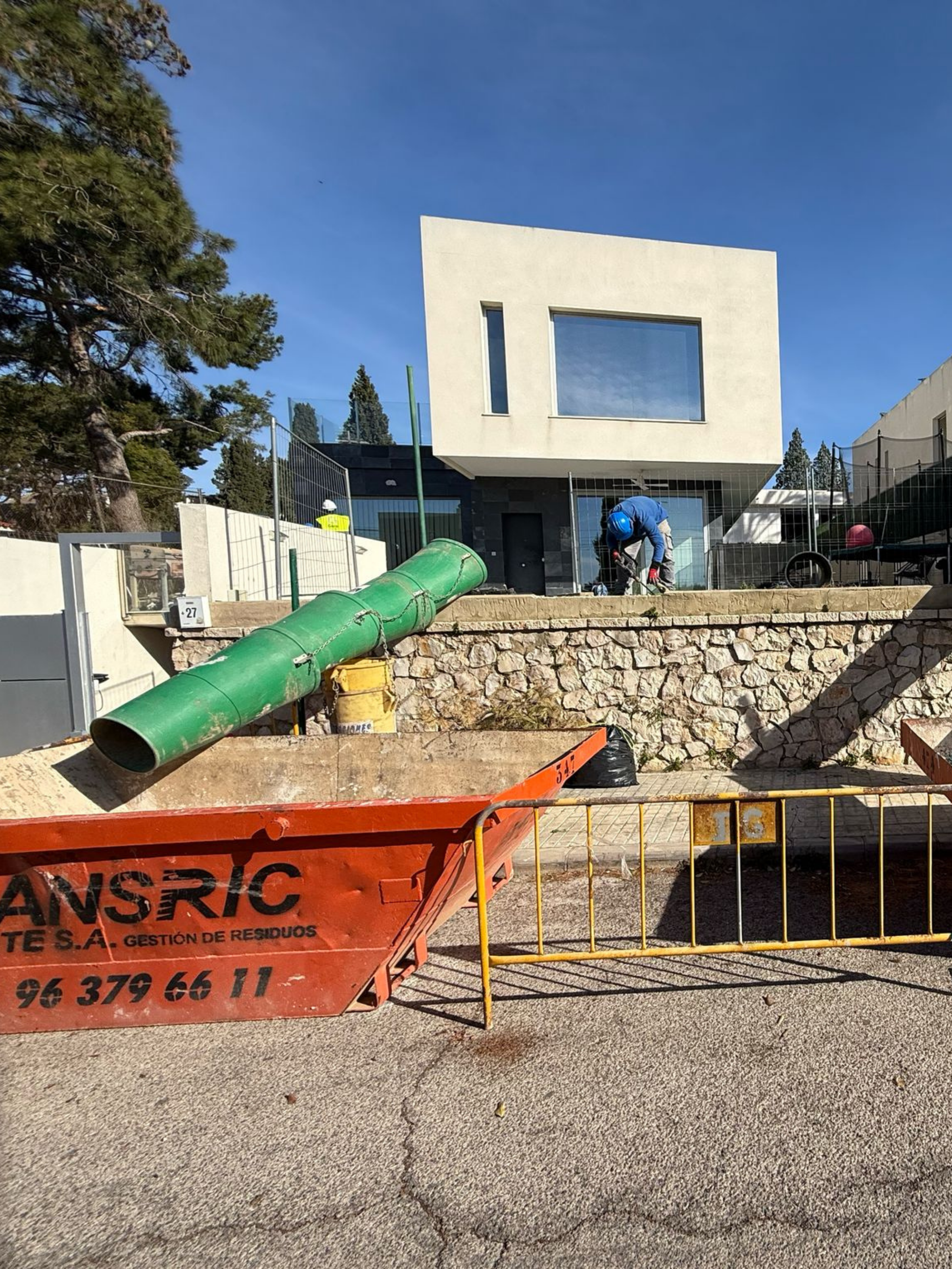 Vivienda unifamiliar de diseño moderno en construcción, con operarios en obra, contenedor de escombros y sistema de bajada de residuos instalado.