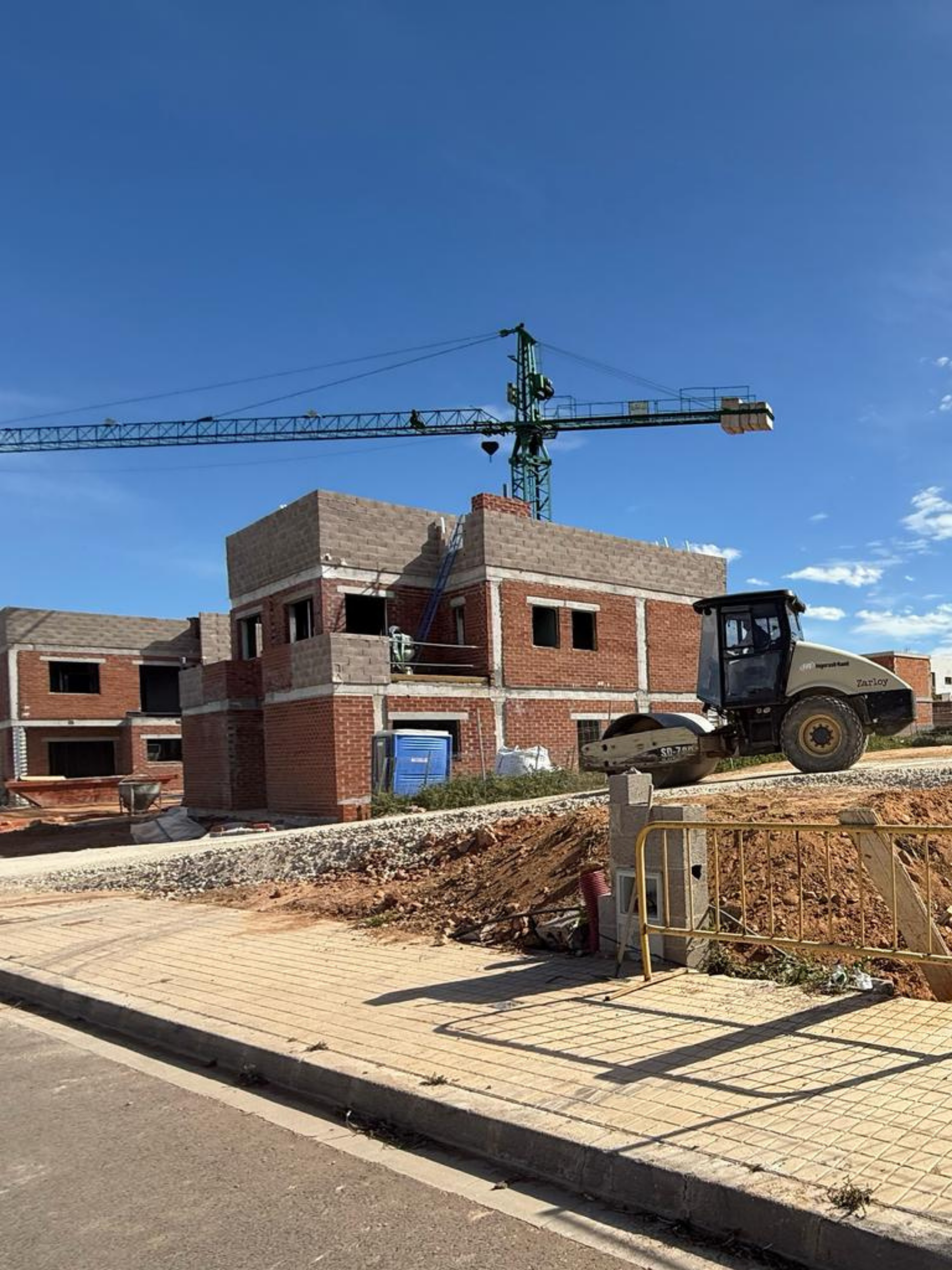 Conjunto de viviendas unifamiliares en fase de obra con estructura de ladrillo visto, grúa torre en funcionamiento y rodillo compactador sobre zona de movimiento de tierras.